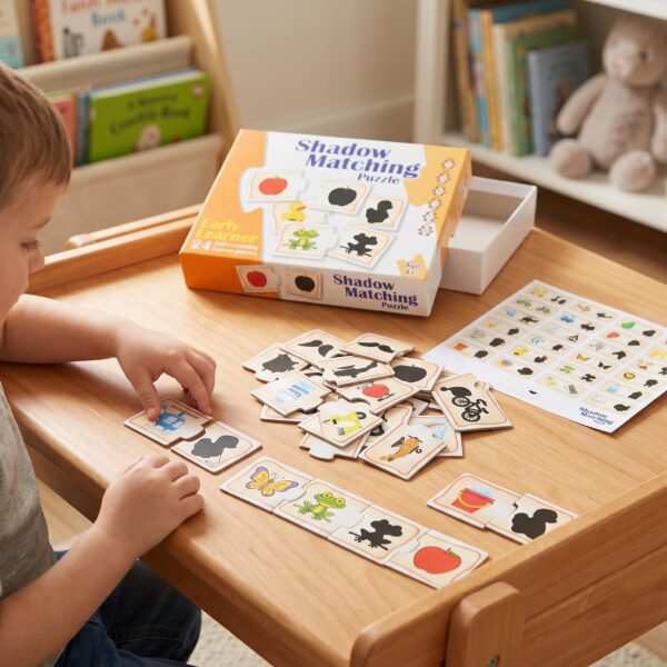 A young boy sitting at a wooden desk, focused on assembling KidNzo Shadow Matching 2-piece puzzles, pairing colorful objects like a frog and helicopter to their silhouettes.
