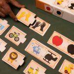 A young boy sitting at a wooden desk, focused on assembling KidNzo Shadow Matching 2-piece puzzles, pairing colorful objects like a frog and helicopter to their silhouettes.