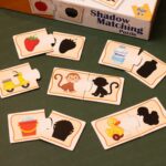 A young boy sitting at a wooden desk, focused on assembling KidNzo Shadow Matching 2-piece puzzles, pairing colorful objects like a frog and helicopter to their silhouettes.